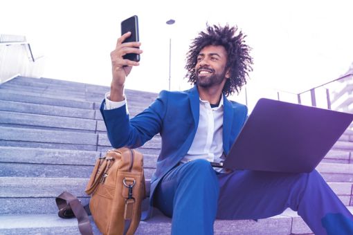 Empresario feliz con laptop tomándose una selfie con su teléfono móvil mientras está sentado en las escaleras.