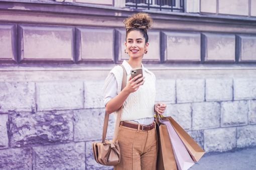 Mujer alegre utilizando su teléfono móvil y llevando bolsas de compras mientras realiza compras en la ciudad