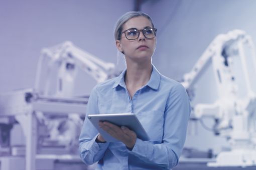 Persona con camisa azul usando una tablet en planta industrial, rodeado de robots automatizados en un ambiente tecnológico avanzado.