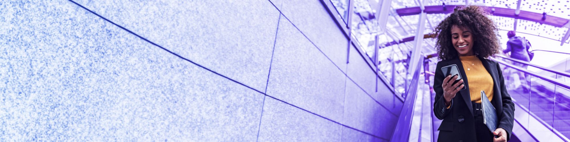 Persona en una escalera mecánica dentro de un edificio moderno. La persona sostiene un teléfono móvil y una carpeta azul. El fondo presenta una estructura de techo de vidrio y paredes grises. Otra persona también está subiendo la escalera mecánica en el fondo. La iluminación natural del día se filtra a través del techo de vidrio, iluminando el espacio interior.