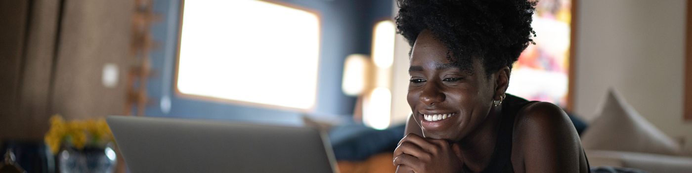 iimagem de uma mulher deita na cama de bruços olhando para a tela de um notebook