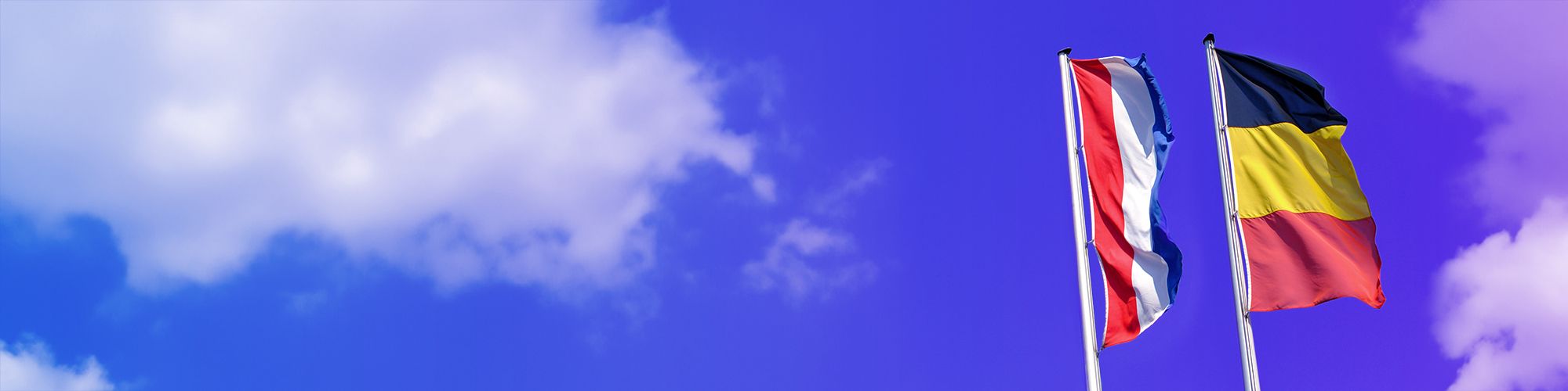Een vlag van België en één van Nederland onder een blauwe hemel met wolken.