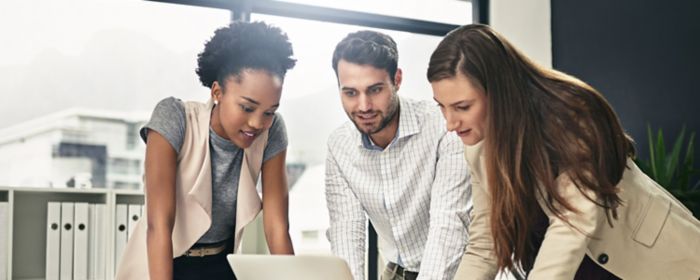 Three people standing near a desk, looking at a laptop.