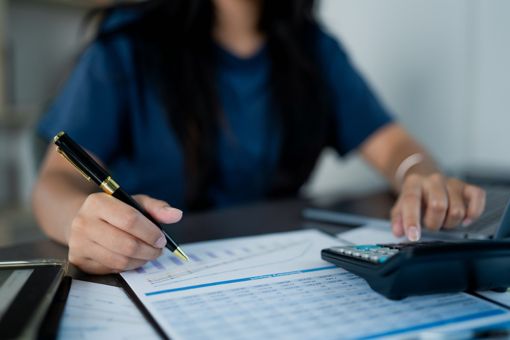 A woman is typing something on a calculator and writing something down on paper.