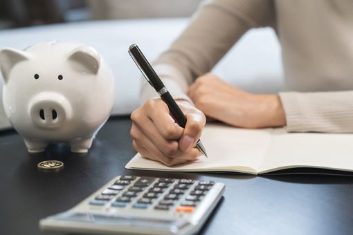Someone is writing something down on paper and there is a calculator and a piggybank on the table