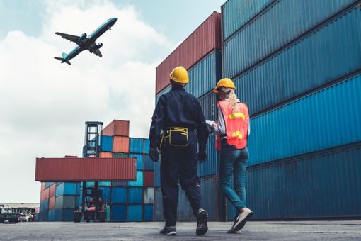 Two people walking, surrounded by containers and a plane flying above them