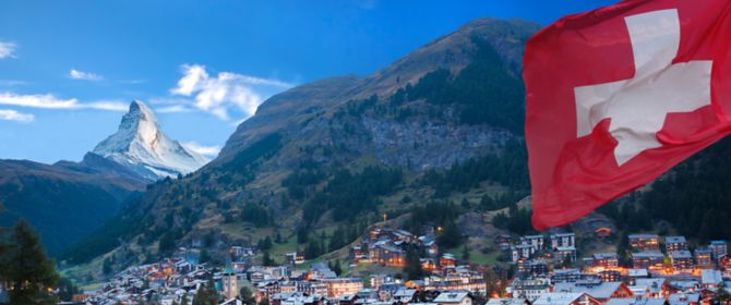 vista de uma cidade, em primeiro plano tem aparece a bandeira da suiça e ao fundo da cidade  montanhas