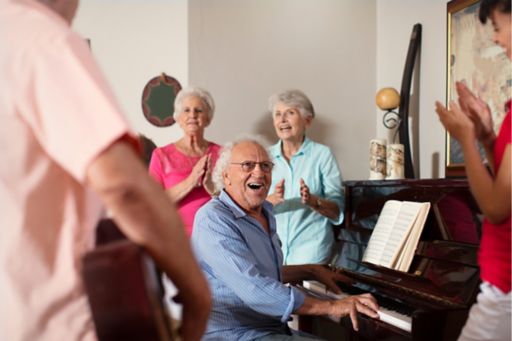 Elderly aged care residents sit around a piano laughing and enjoying music