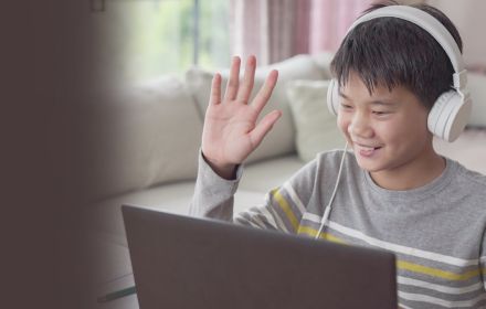 Boy sitting in front of laptop wearing headphones and waving hand