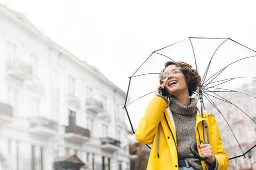Girl holding an umbrella and wearing a yellow jacket is laughing while talking on the phone