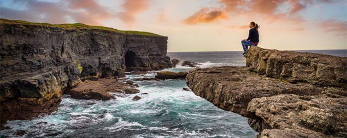 Cliffs in Kilkee at sunset, Ireland. Woman sitting on the edge of a cliff by the ocean.