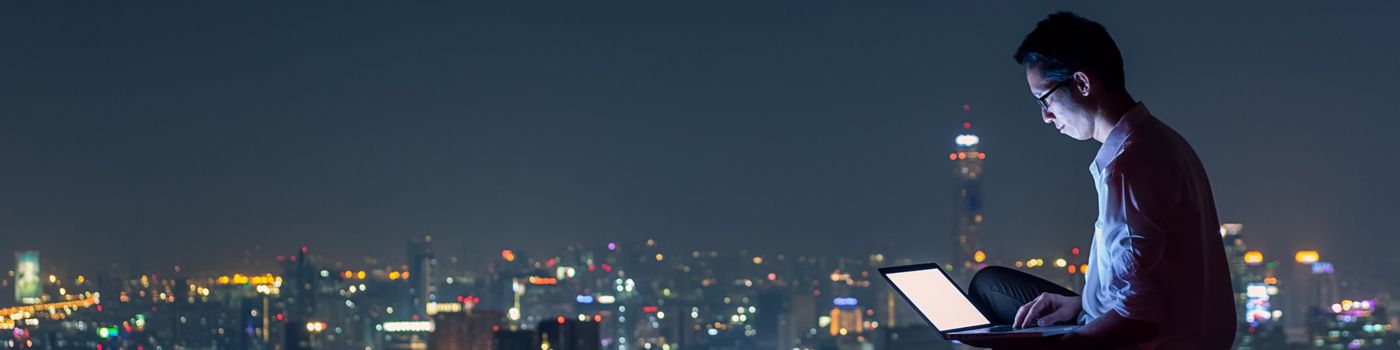 Man working on laptop late night sitting at roof, city lights background