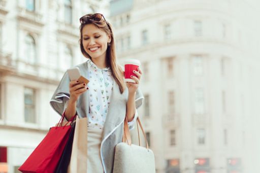 Cheerful woman is shopping in the city. She is holding a coffee to go and using her smart phone.