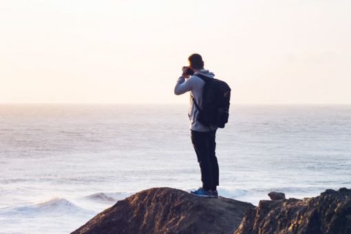 Mann står på et berg ved sjøen og tar bilder med et fotoapparat 