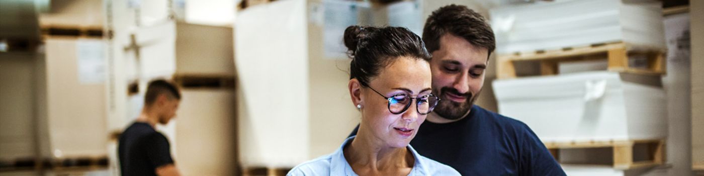 Mann und Frau stehen in Lagerhalle und schauen ein iPad und unterhalten sich