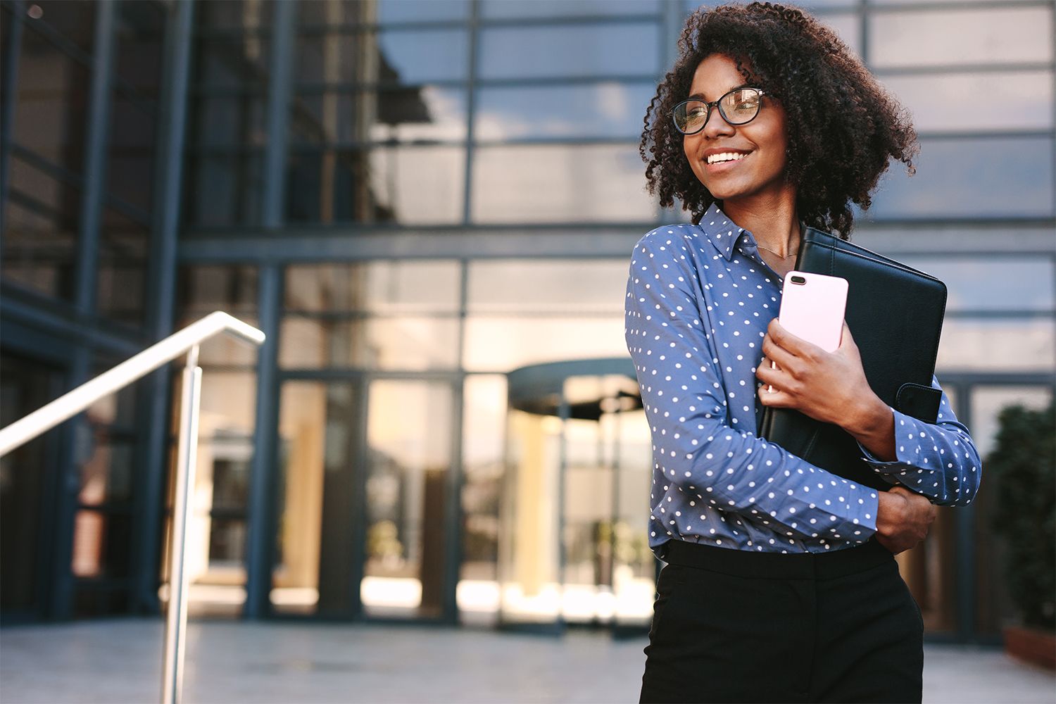 Woman with mobile phone and file standing outdoors and looking away. Business woman standing outside a office building.