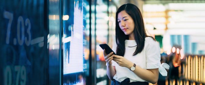 Young Asian businesswoman checking financial trading data on smartphone by the stock exchange market display