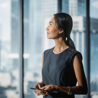 A woman standing beside floor-to-ceiling windows and holding a smartphone, with an urban skyline visible in the background
