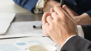A hand of a man is holding a pen and some papers are visible in the background