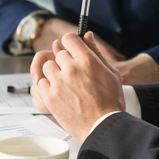 A hand of a man is holding a pen and some papers are visible in the background