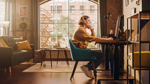Young Handsome Man Working from Home on Desktop Computer in Sunny Stylish Loft Apartment. Creative Designer Wearing Cozy Yellow Sweater and Headphones. Urban City View from Big Window.
