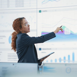 Hispanic Female Senior Data Scientist Reviewing Reports Of Risk Management Department On Big Digital Screen In Monitoring Room. Diverse Consulting Company Employees Working Behind Desktop Computers.