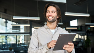 Smiling busy young European business man company employee using tablet standing at work. Happy male professional executive manager business owner using tab computer device looking away in office.