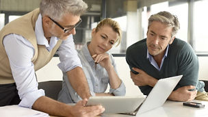 Three businesspeople look at a tablet and laptop together.
