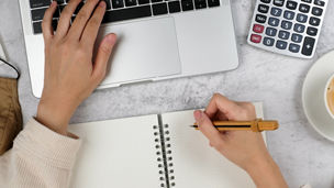 Someone is writing something down in a notebook, and typing something on a computer with the other hand.