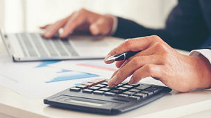 Deux mains, dont l'une tape sur un ordinateur portable et l'autre tape quelque chose sur une calculatrice tout en tenant un stylo