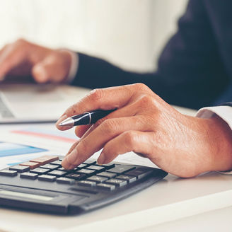 Two hands, one typing on a laptop and the other typing something on a calculator while holding a pen.