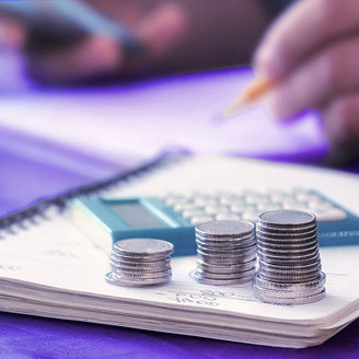 A man is sitting at a desk, holding his phone and writing something down on a paper on which coins are laying