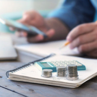A man is sitting at a desk, holding his phone and writing something down on a paper on which coins are laying