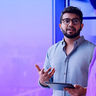 Un professionnel portant des lunettes, tenant une tablette, fait des gestes pendant une discussion. Il se tient devant une grande fenêtre avec une ambiance technologique.