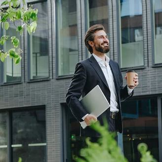 Man walking in front the building while holding a cup of coffee and laptop