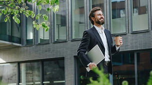 Man walking in front the building while holding a cup of coffee and laptop