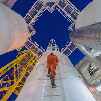 Worker in an orange protective suit climbing a vertical ladder between large industrial pipes and structures at an outdoor facility during nighttime