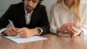 Een man die een papier ondertekent en een vrouw die een ring rond haar vinger doet zitten naast elkaar aan tafel.