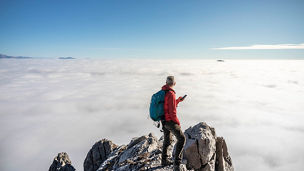 Hiker on top of the mountain with blue clear sky over clouds using smart phone