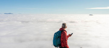 Hiker on top of the mountain with blue clear sky over clouds using smart phone