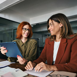 Professional female coworkers employees looking at digital tablet discussing corporate project sharing ideas at work. Two busy happy business women talking using tab working together at desk in office