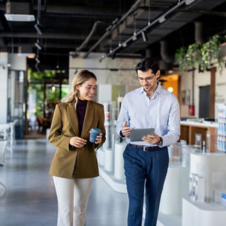 Two young professionals walking through office discussing work with tablet and coffee