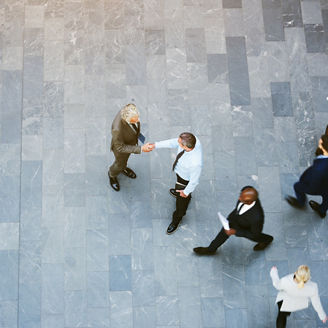 From above two adult business men shaking hands met in office hall with workers walking around.