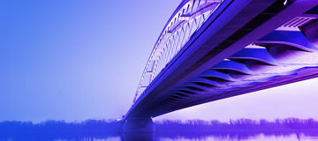 Eine moderne Brücke, die sich über einen Fluss erstreckt, mit einer futuristischen Architektur. Die Brücke ist in einem blauen und violetten Farbton beleuchtet, und der Himmel sowie das Wasser spiegeln diese Farben wider.