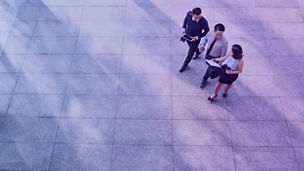 Three business professionals walking together on a large tiled floor, holding documents and a tablet.