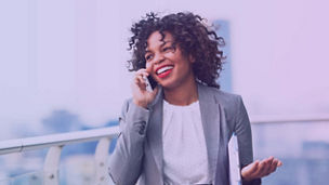 Mulher de negócios sorridente ao telefone num terraço exterior, com paisagem urbana ao fundo.