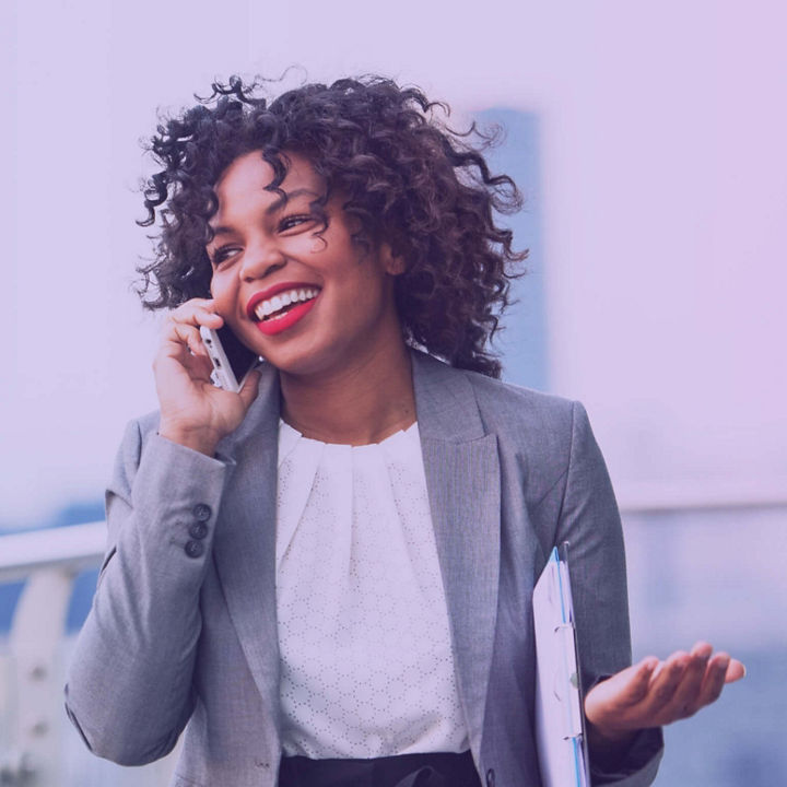 A smiling businesswoman on the phone on an outdoor terrace, with a cityscape in the background.