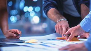 Mãos de três profissionais sobre uma mesa de trabalho, a organizar documentos impressos e notas adesivas coloridas ao lado de um computador portátil.