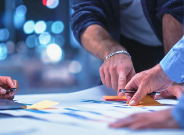 Mãos de três profissionais sobre uma mesa de trabalho, a organizar documentos impressos e notas adesivas coloridas ao lado de um computador portátil.