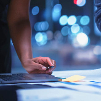 Mãos de três profissionais sobre uma mesa de trabalho, a organizar documentos impressos e notas adesivas coloridas ao lado de um computador portátil.